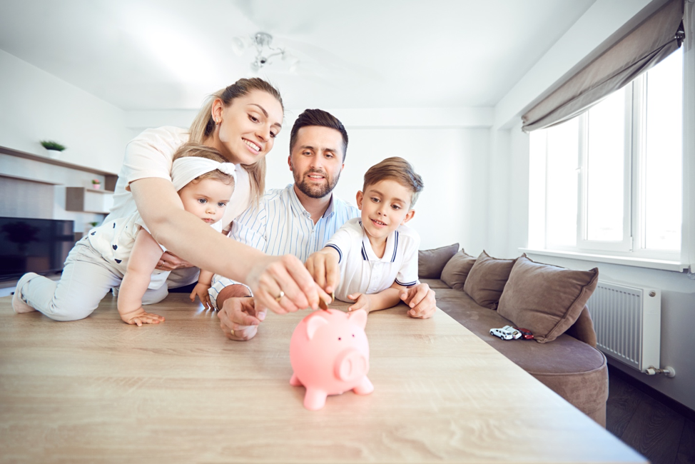 Mother, father, daughter and son sit around the kitchen table and put a coin in a pink piggy bank